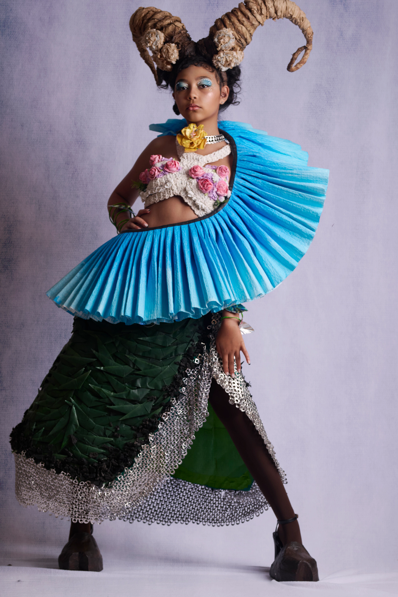 Recycle the Runway: Veyra A student models an avant-garde outfit made from recycled materials. The outfit features a skirt made from green origami and soda can tabs, a large blue ruffle, a crochet bodice, a choker made from aluminum soda cans, and large goat horns.