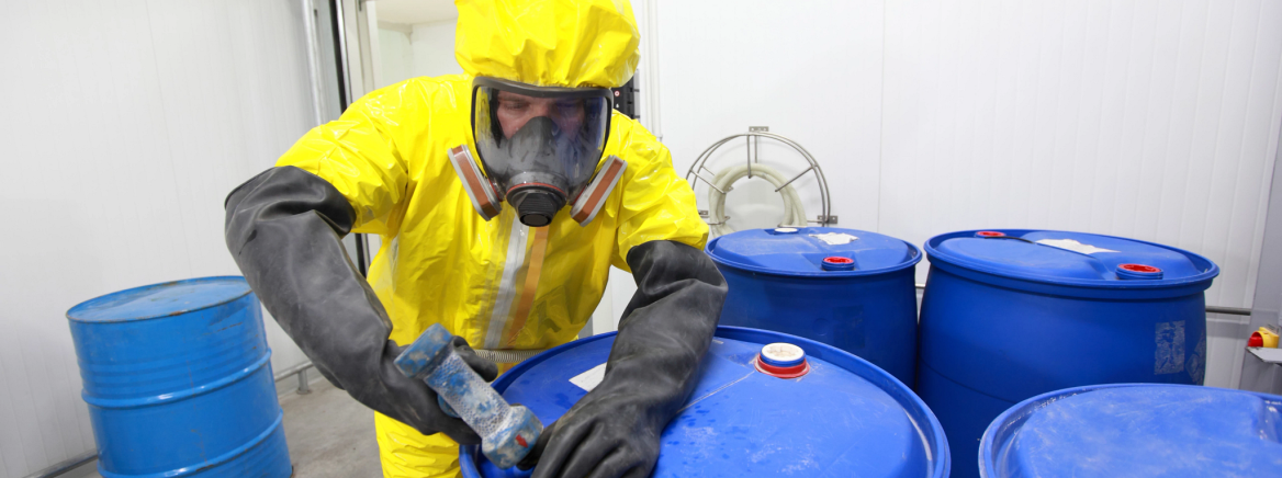man in protective suit handling blue barrels of industrial waste